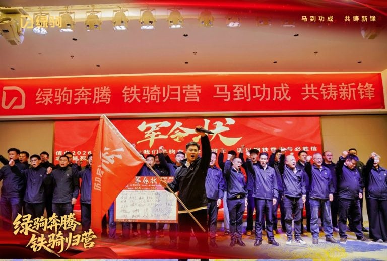 Group of people on stage during the 2026 Green Steed Rally, celebrating teamwork and dedication with a leader holding a flag, in front of a red banner with Chinese characters.