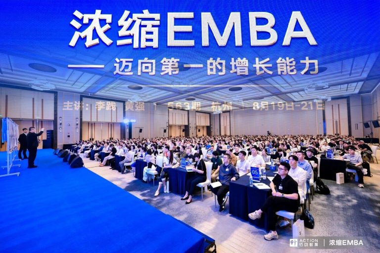 Large conference hall hosting EMBA event with many attendees seated at tables, stage with speakers, and prominent blue branding for EMBA program.